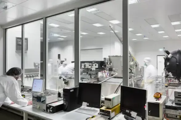 Blick in zwei Reinräume, die durch eine Serie großer Glasfenster voneinander abgetrennt sind. Im Vordergrund Labortische, auf denen Monitore, Tastaturen und Rechnergehäuse stehen. Eine Person in Reinraumkleidung ist von der Kamera weg gewandt und bedient nach vorne gelehnt stehend einen Computer. Im Hintergrund mehrere Laborbänke mit vielfältiger Ausrüstung, an der zwei weitere Personen arbeiten.