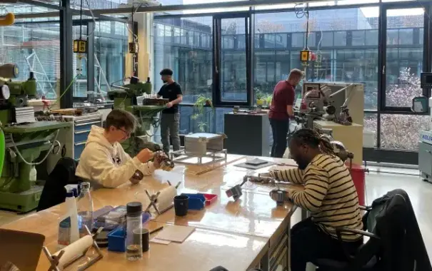 Blick in eine großräumige lichtdurchflutete Werkstatt. An einem aus mehreren Tischen zusammengestellten großen Arbeitsbereich in der Mitte sitzen zwei junge Menschen über ihre Projekte gebeugt. Im Hintergrund stehen zwei weitere Menschen an feinmechanischen Geräten und bedienen diese. Durch die Fensterwand im Hintergrund öffnet sich die Sicht auf einen Innenhof.