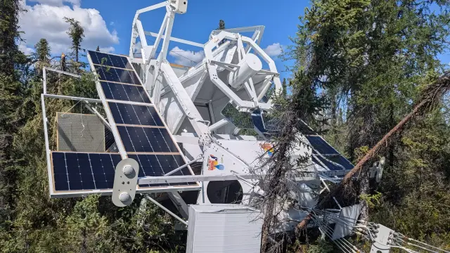 Inmitten einer Umgebung mit Bäumen hängt schief eine große Struktur mit vielen weißen Streben und daran seitlich angeordneten teilweise abgebrochenen Solarpanelen: das gelandete Sunrise III Teleskop. Die Bäume sind grüne Nadelbäume, die Baumstämme sind teilweise umgeknickt. Es ist ein sonniger Tag mit blauem Himmel und wenigen wießen Wolekn im Hintergrund. Die Sonne scheitn so hell, das das Weiß von Teleskop und Gondel intensiv leuchtet, während die Solarpanele dunkel schimmern.