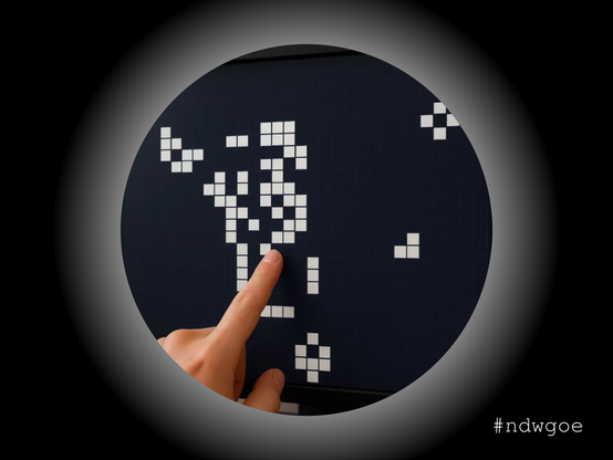 Analoge Version des Game of Life in weiß auf schwarzer Tafel mit einen zeigenenden Finger davor durch ein Guckloch betrachtet