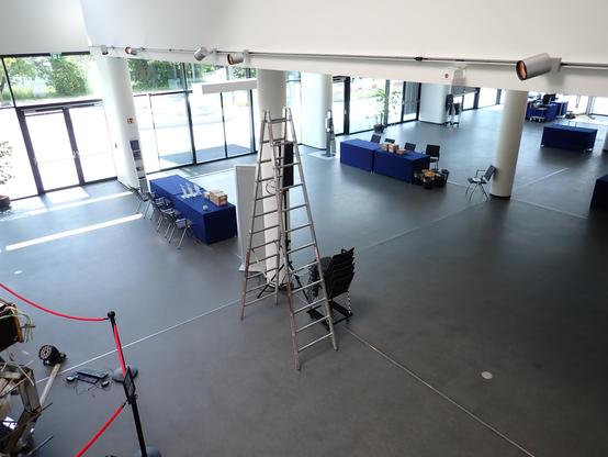 A picture taken from an elevated viewpoint from inside a building looking back towards the building's entrance area. There are tables to both sides of a closed large glass entrance door and chairs arranged behind the tables that face the corridor created by the tables. Closer to the image front plane, there is a large ladder next to a staple of additional chairs. Off in the far background to the right, more tables mark the areas where stands are being set up. In the front left image corner, red…