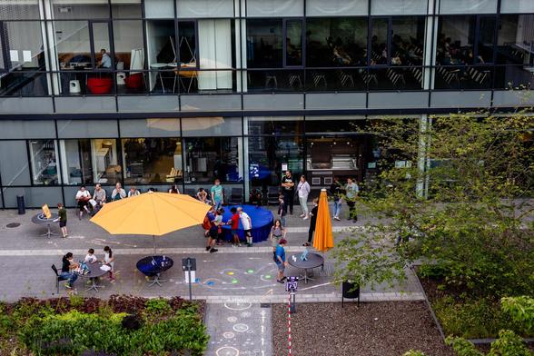 A wide-angle view of a courtyard, framed by two stories of its host buildung. In the courtyard, chalk drawings on the floor, a parasol, and a round blue cloth. Little people scurrying around it all.