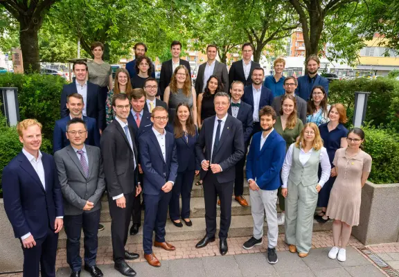 Otto Hahn medal awardees group picture at the award ceremony: More than two dozen young people on four levels of steep stair steps in formal attire all facing the camera, with a green garden in the background.