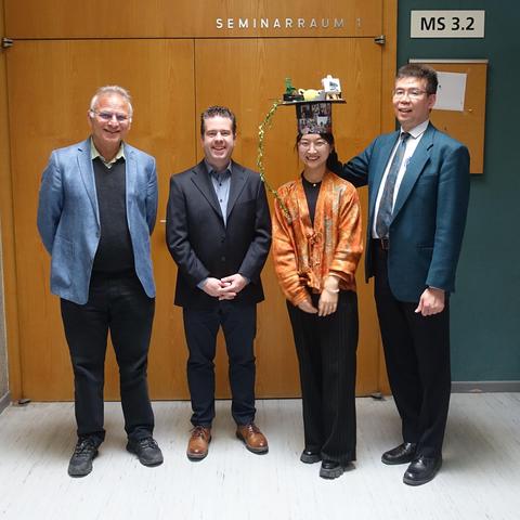 Four people standing next to each other on front of a wood-panelled wall in formal clothing: from left to right, Sami Solanki, Ferdinand Plaschke, Ziwen Huang, Yasuhito Narita. Ziwen is wearing a creatively decorated doctoral hat. All four smile and look happy.