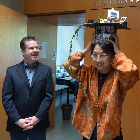 Ziwen putting on her doctoral hat: both hands on the hat, trying to balance it. Ferdinand is standing next to her and watching, smiling.
