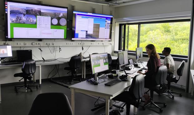 Two scientists are seated at tables in a lab room with three small monitors in front of them and two large screens mounted on the wall. The large monitors show various monitoring images of a telescope and the data it is taking.