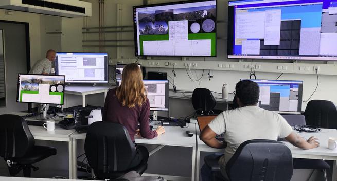 A full view of the operations center again, from a different angle as before and this time showing the backs of three busy scientists facing various monitors both directly in front of them as well as the two large screens on the wall of the room. The scientist in the middle is busy using her mouse clicking on things, and her upright posture suggests a high level of concentration.