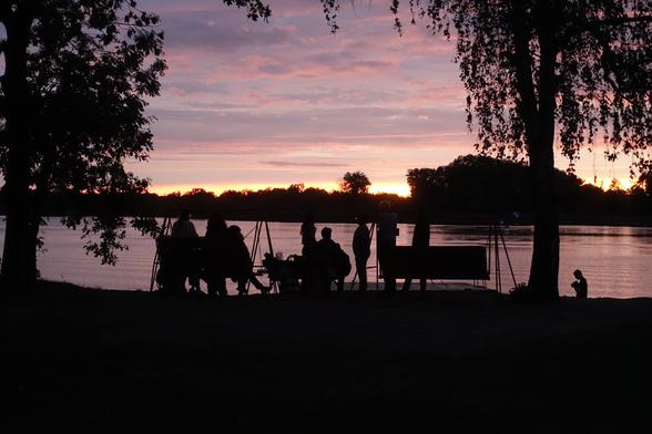 An evening scene by the lake side: Colors are all subdued pinks and blues, with dark silhouettes. Sitting and standing on an elevated lake shore, a group of people only visible as silhouettes against the still reflecting surface of the lake and a dramatic sky above the treeline on the opposite shore.