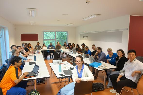 A seminar room with a group of roughly thirty young people sitting around a U-shaped confernce table, looking up at the camera.