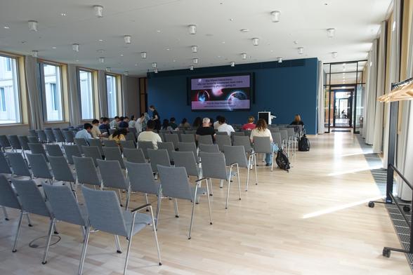 View from the back of a large modern seminar room to the front of the room with the title slide of an exoplanets talk projected. A group of people are sitting on chairs in the room, anticipating the start of the talk.