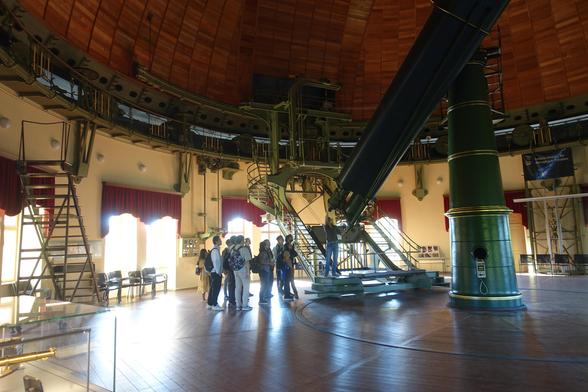 Inside a large cupola with a wooden roof, a group of people are looking at a large refractor telescope. It's two very long tubes on a mount, painted dark green. The instrument is huge in comparison to the people standing underneath it.