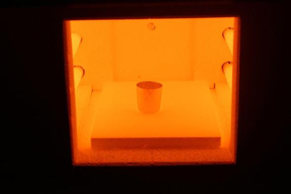 A sample in a cup in a furnace, with the scene glowing hot red behind the protective transparent door.