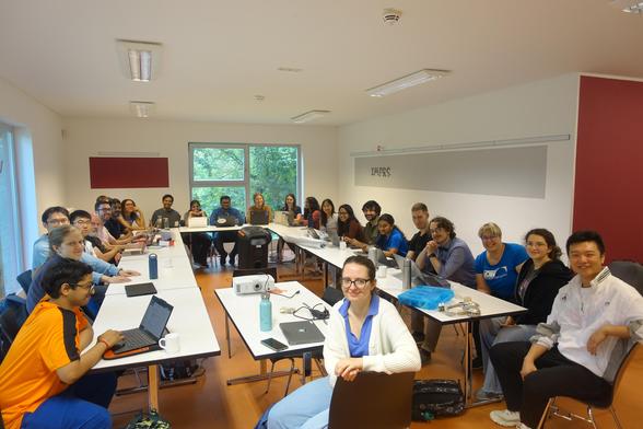 A seminar room with a group of roughly thirty young people sitting around a U-shaped conference table, looking up at the camera.