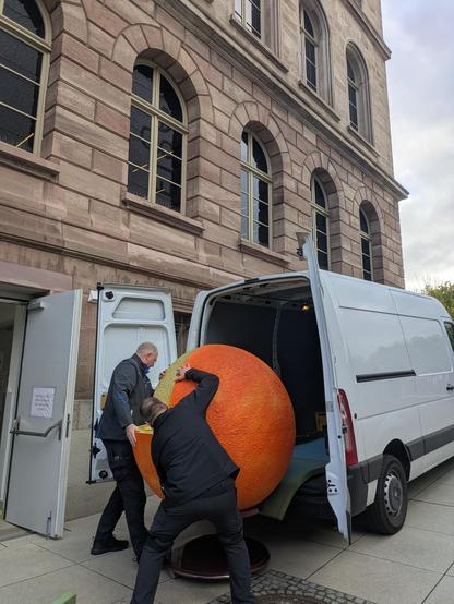 Two people pushing a large model of the Sun into a van. In the background: the Forum Wissen.