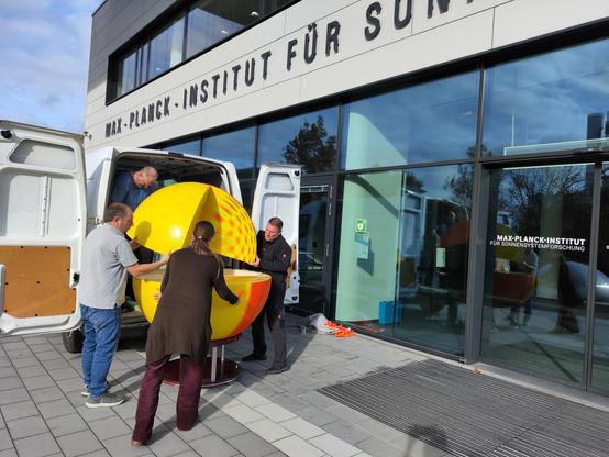 Four people lifting a large model of the Sun out of a white van. In the background: main entrance of the Max Planck Institute for Solar System Research.