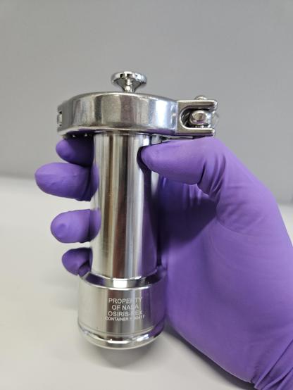 Hand in purple lab glove holds a cyclindric, silvery sample container with the label "PROPERTY OF NASA - OSIRIS-Rex".