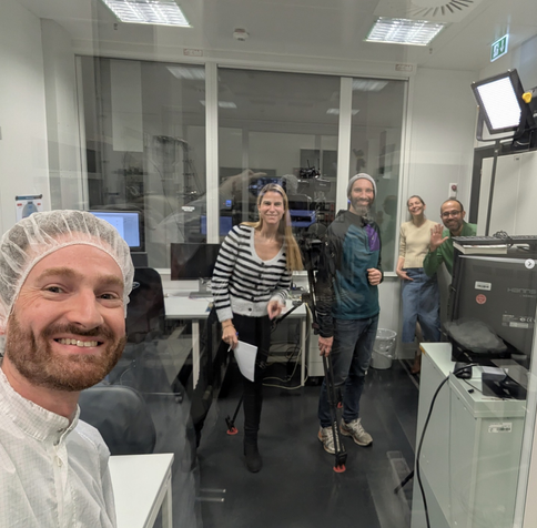 Ein Kamerateam sowie MPS Mitarbeiter*innen in der Nähe des Reinraums. Eine Person in Reinraumkleidung schaut von links unten ganz nah ins Bild, sie macht ein Selfie.