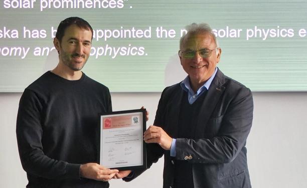 Two people present a framed certificate in front of a screen displaying text.