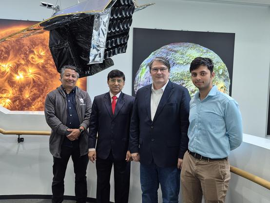 Four men in formal attire standing next to each other, all looking towards the camera with serious yet friendly expressions. Above their heads, a large satellite model hangs from the ceiling. Further in the background, parts of two large wall murals depicting the Sun and a minor planet are visible.
