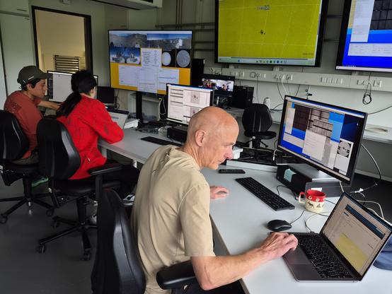 Operations center with 3 scientists working at desks and several large monitors showing the GREGOR telescope, a sunspot and a full-disc views of the Sun. 