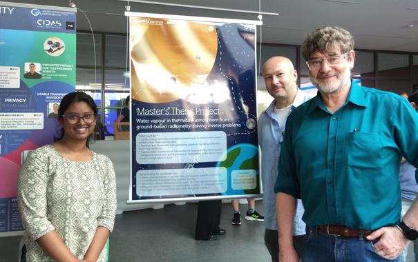 A young woman standing to the left of a poster, two men standing yo the right. The poster has a planet featuring prominently in the upper half and text below.