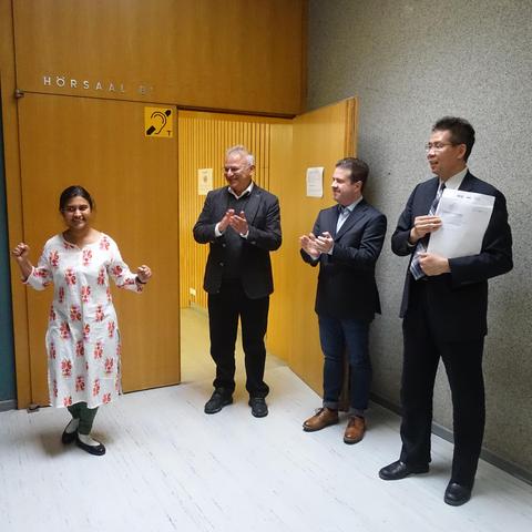 Young woman just emerged through a wooden door doing a little happy dance. To her left, three man clapping. This is the moment the new doctor is presented to the world by her committee members.