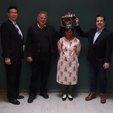 Formal picture of a new doctor with her committee members. The successful candidate is wearing a doctoral hat, held on her head by the two people to her left an right. A forth person stands further to her right. All are in formal attire and looking towards the camera.