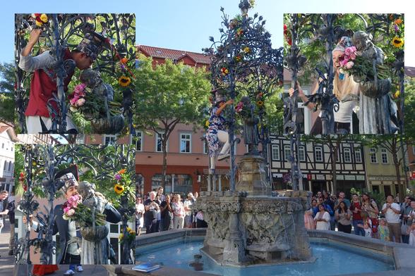 Composite showing a fountain with a goose girl statue on a high pedestal surrounded by a crown on a sunny day. A woman wearing a tall doctoral hat has climbed the struture and is leaning towards the statue. Three inset images show close-ups of different people all also kissing the statue.
