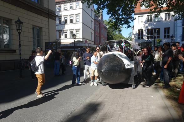 A doctoral cart in the shape of a space shuttle seen from the front, with people in it, and people outside pushing and steering it onto a city road. Even more people all around point cameras at the scene.