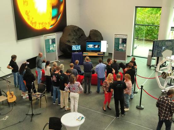 A setup oft large monitors in the corner of a high-ceilinged building. The ESA live stream is playing on the central monitor. To both sides there are BepiColombo posters. A crowd is seen from behind, watching.
