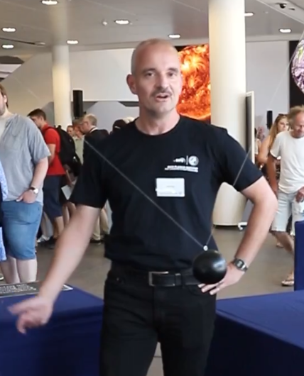 A man in a black shirt standing and explaining an experiment involving a pendulum. The pendulum is visible right in front of the speaker, swinging back and forth. To the right, a monitor with a star whose brightness changes. In the background, a large foyer swarming with people.